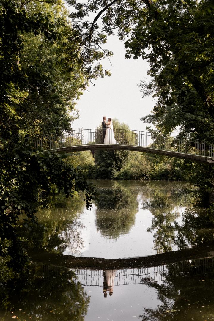Brautpaar steht romantisch auf einer Brücke über einem ruhigen Teich, eingerahmt von üppigem Grün, mit Spiegelung im Wasser.