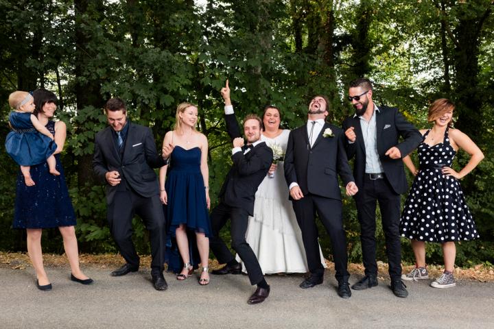 Brautpaar posiert für ein Gruppenfoto mit Gästen in fröhlicher und ausgelassener Stimmung vor einer grünen Waldkulisse.